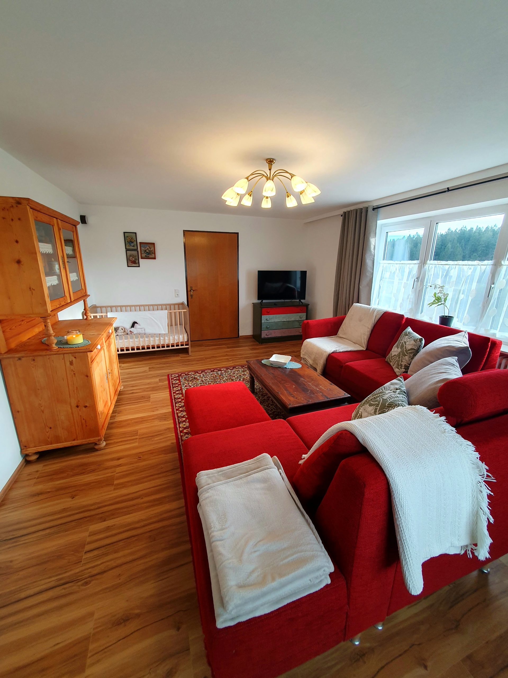 Wohnzimmer mit rotem Sofa, Gitterbett und Blick in den Wald